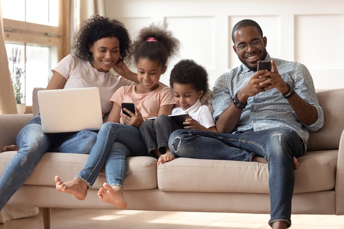 A family of four seated on a couch, each engaged with their own wireless device.