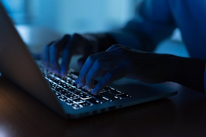 A hacker typing on a backlit laptop keyboard in a dimly-lit room. 
