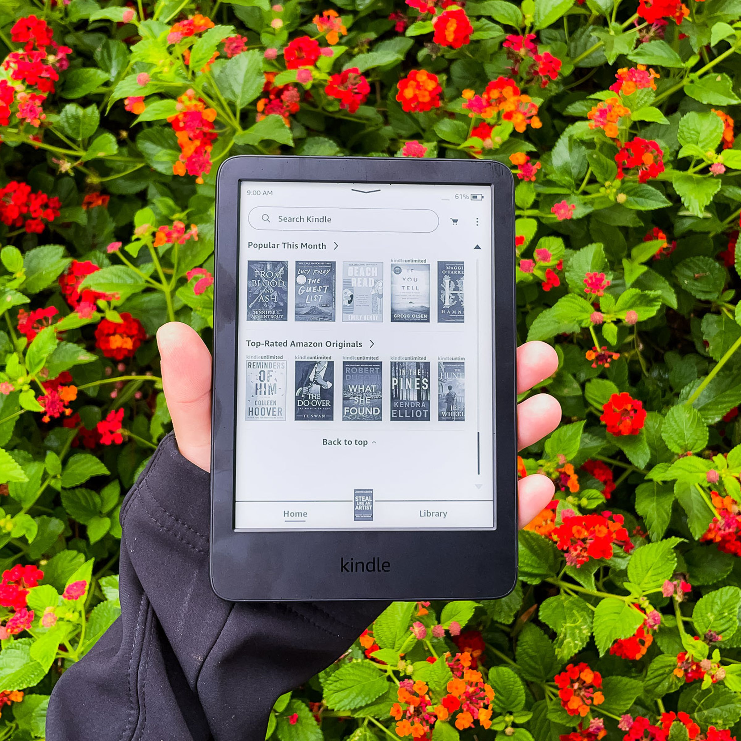 A hand holding the 2022 Kindle in front of red flowers.