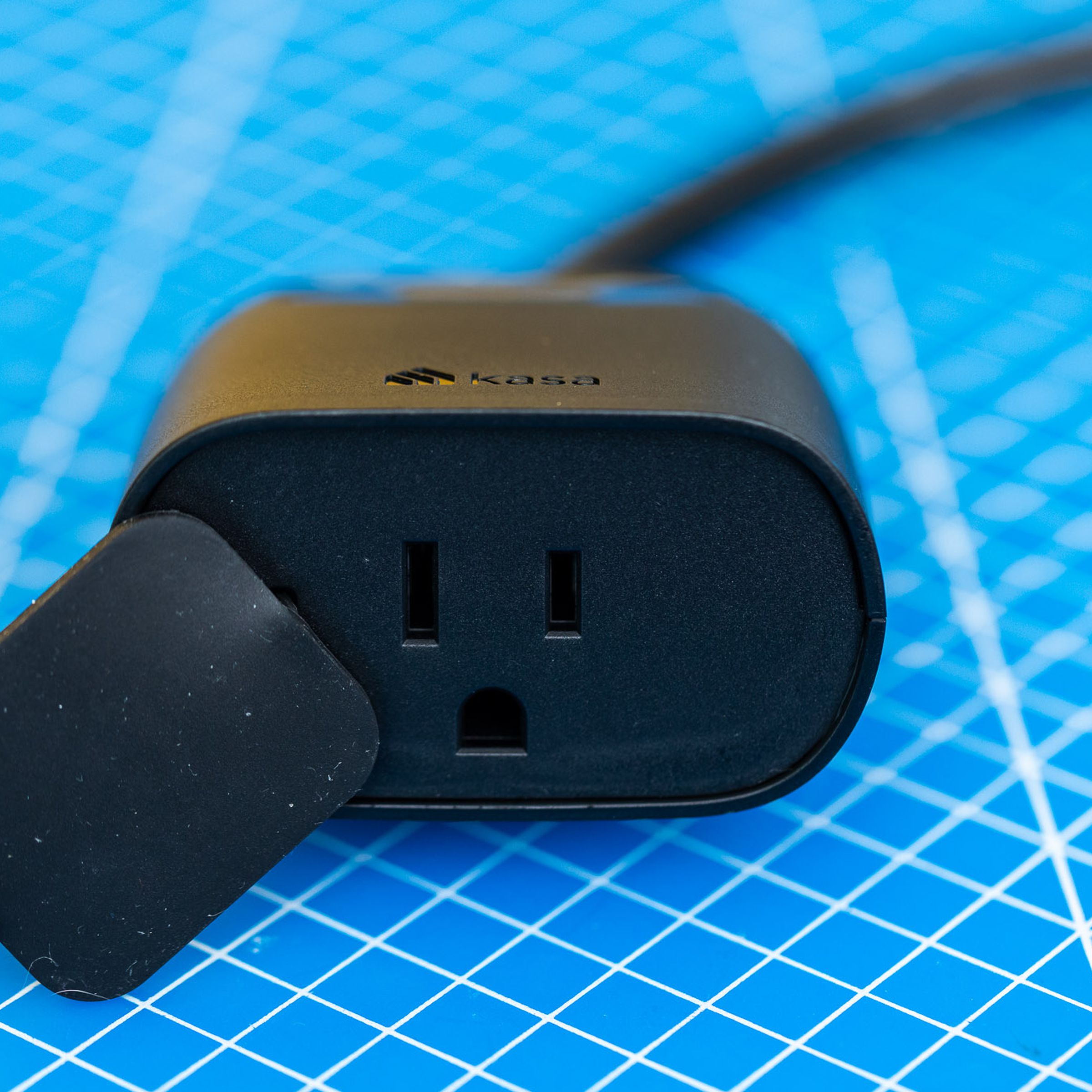 A black, two-outlet outdoor smart plug. It has a short cable leading to its own power plug, and a flap to protect the outlets from moisture. It is on a blue background with a grid of white lines.