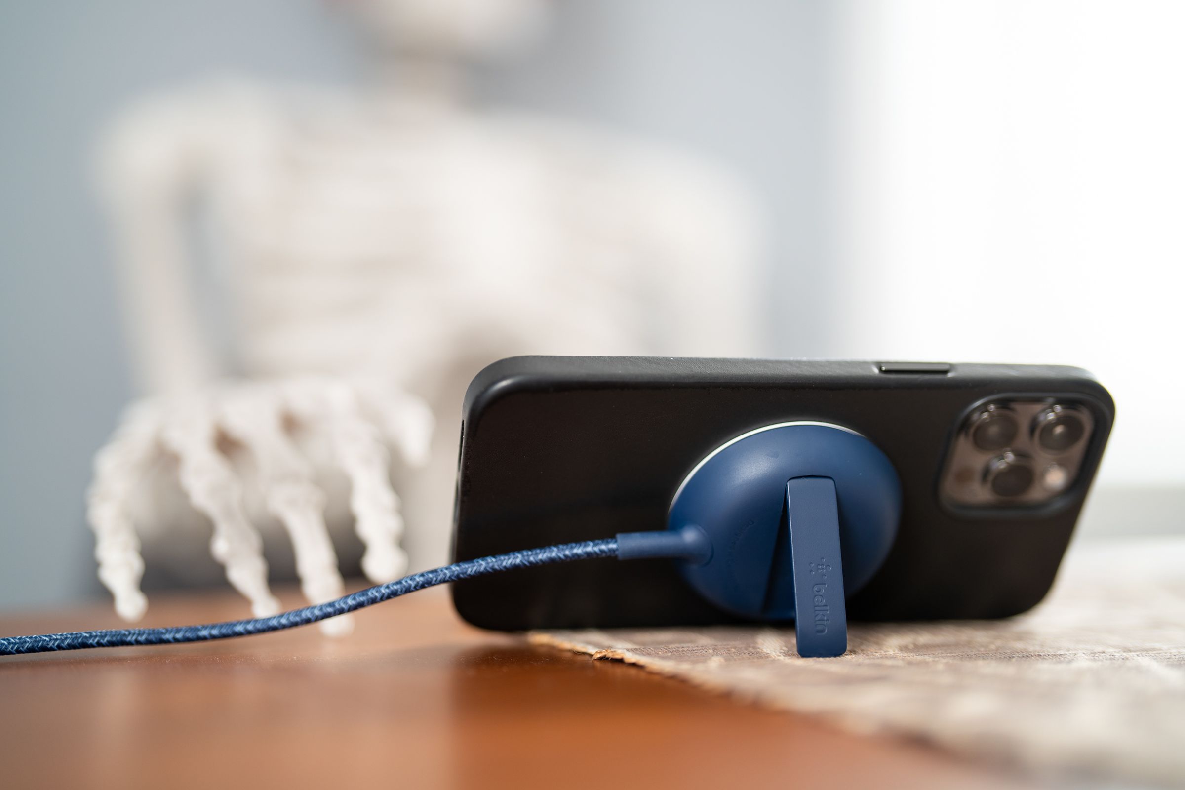 An iPhone propped up horizontally by the kickstand of a Belkin MagSafe charger on a table with a prop skeleton sitting in a chair and looking at the phone.