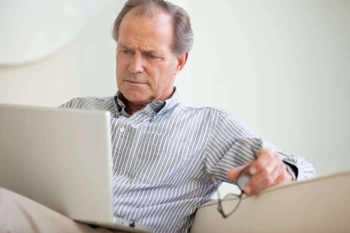 A person holding reading glasses in their right hand while critically reading content from an open laptop.