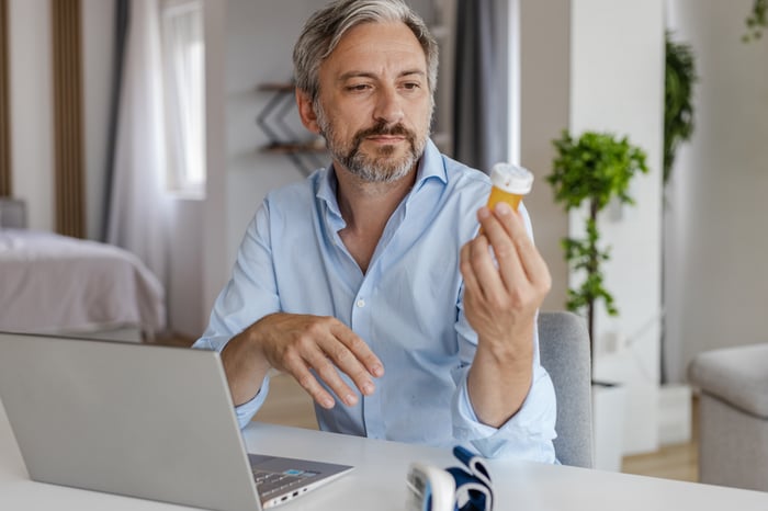 A person at a laptop holding a bottle of pills.