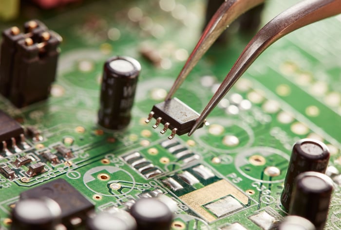 A pair of tweezers holding a computer chip above a circuit board.